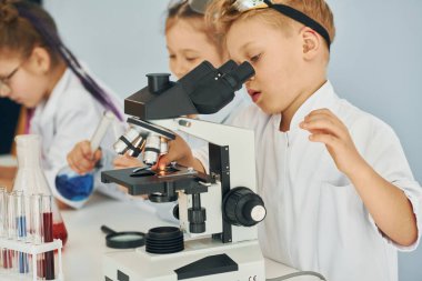 Mikroskop kullanarak. Beyaz önlüklü çocuklar laboratuarda ekipman kullanarak bilim adamlarını oynuyorlar..