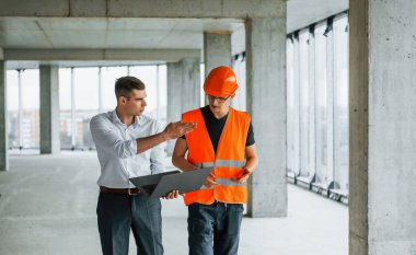 İşlemi planlıyorum. Takım elbiseli bir adam ve turuncu giysili bir tamirci inşaatta çalışıyor..