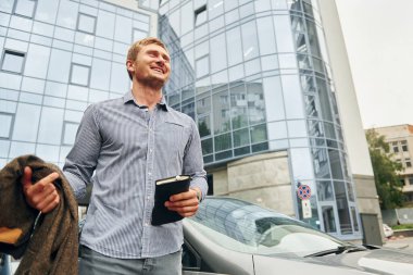 Elinde not defteriyle. Gündüz vakti şehirde gezen bir adam..