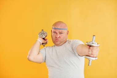 Dambıl kullanarak alıştırma yapıyor. Spor kravatlı komik şişman adam sarı arka plana karşı..