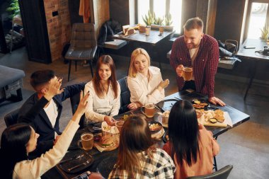 Adam ayakta ve gülümsüyor. Bir grup genç arkadaş barda birlikte oturup bira içiyorlar..