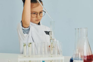 Test tüplerinde bulunan sıvıyla çalışıyor. Palto giyen küçük bir kız laboratuarda ekipman kullanarak bilim adamını oynuyor..