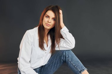 Beyaz gömlekli ve kot pantolonlu. Genç ve güzel bir kadın stüdyoda kameraya poz veriyor..