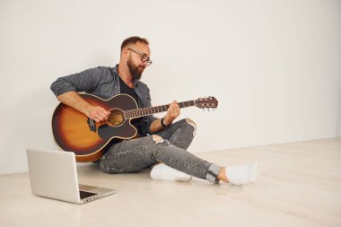 Dizüstü bilgisayarla. Gündelik giysiler içinde ve akustik gitarlı bir adam içeride..
