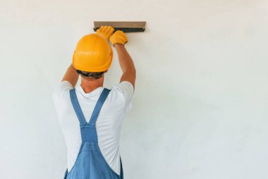 Beyaz duvarın yanında duruyor. Gündüz vakti inşaatta üniformalı genç bir adam çalışıyor..