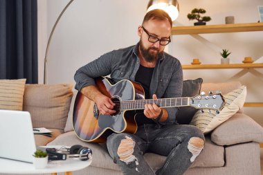 Kanepede. Akustik gitar çalıyor. Sıradan elbiseli adam içeride..