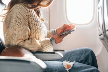 Sitting with phone in hands. Young female passanger in casual clothes is in the plane. clipart