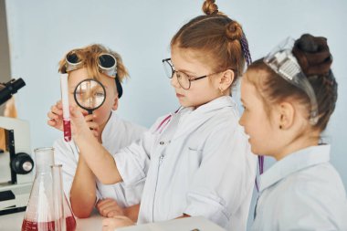 Test tüpleriyle masanın yanında duruyor. Beyaz önlüklü çocuklar laboratuarda ekipman kullanarak bilim adamlarını oynuyorlar..