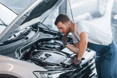 Kaput açıldı. Beyaz gömlekli ve mavi üniformalı genç adam otomobil tamir ediyor..