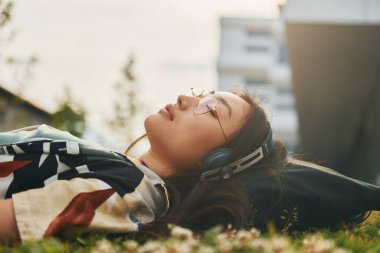 Çimlerin üzerinde dinleniyor. Kulaklıkla. Genç Asyalı kadın gündüz vakti dışarıda..