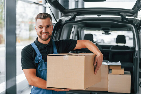 Holding box. Delivery man in uniform is indoors with car and with order.