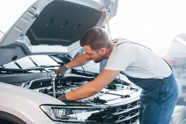 Quality service. Young man in white shirt and blue uniform repairs automobile.