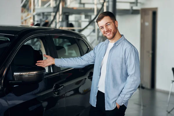 Successful man in glasses standing near brand new car indoors.