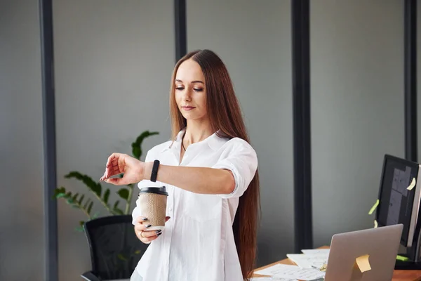 With coffee. Young adult woman in formal clothes is indoors in the office.