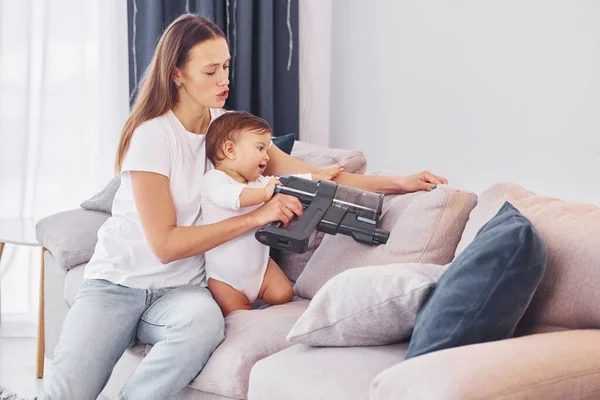 坐在床上 用吸尘器吸尘 妈妈和她的小女儿一起呆在家里 — 图库照片