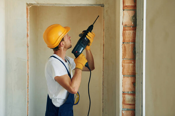 Holding electric drill in hands. Young man working in uniform at construction at daytime.