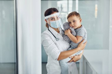 Elinde küçük bir çocukla. Klinikte beyaz önlüklü profesyonel doktor var..