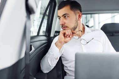 Dizüstü bilgisayarla çalışıyor. Beyaz gömlekli genç adam modern bir otomobilin içinde oturuyor..