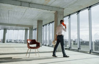 Telefon kullanarak sohbet ediyorlar. Resmi kıyafetli genç adam içeride inşaatta çalışıyor..