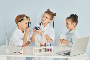Test tüpleriyle masanın yanında duruyor. Beyaz önlüklü çocuklar laboratuarda ekipman kullanarak bilim adamlarını oynuyorlar..