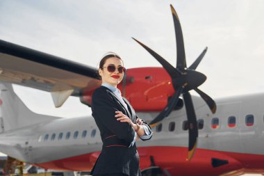 Resmi siyah elbiseli genç hostes uçağın yanında dikiliyor..