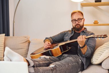 Kanepede. Akustik gitar çalıyor. Sıradan elbiseli adam içeride..