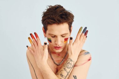 Genç eşcinsel adam stüdyoda duruyor ve kameraya poz veriyor. Gökkuşağı makyajlı tişörtsüz adam..