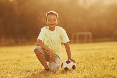 Futbol topuyla oturuyor. Afrikalı Amerikalı çocuk yaz günlerinde tarlada eğleniyor..