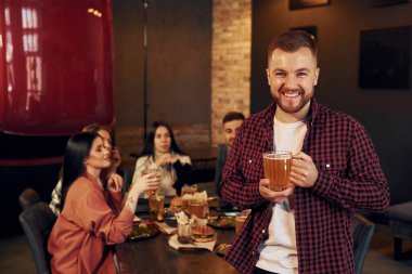 Adam ayakta ve gülümsüyor. Bir grup genç arkadaş barda birlikte oturup bira içiyorlar..