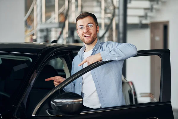 Successful man in glasses standing near brand new car indoors.