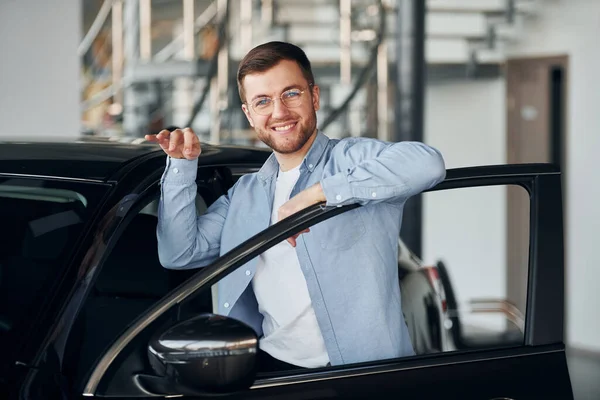 Kapalı alanda yepyeni bir arabanın yanında duran gözlüklü başarılı adam..