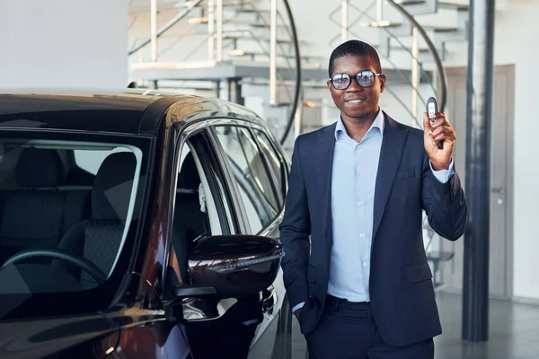 Holding keys in hands. Young african american businessman in black suit is the autosalon.