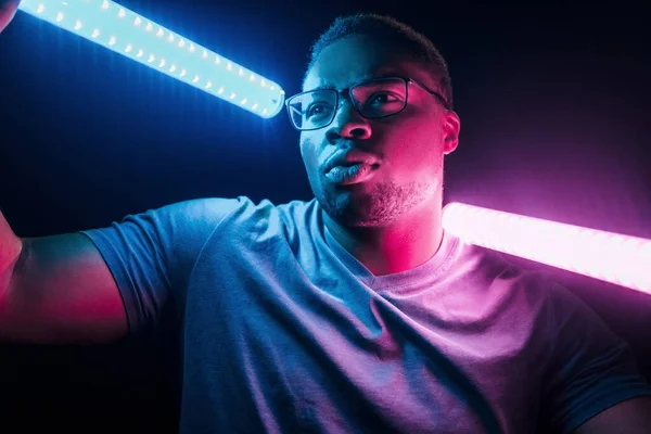 Holds lighting equipment. Futuristic neon lighting. Young african american man in the studio.