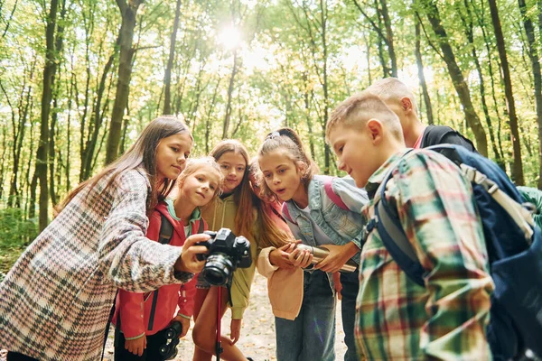 Holding camera. Kids in green forest at summer daytime together.