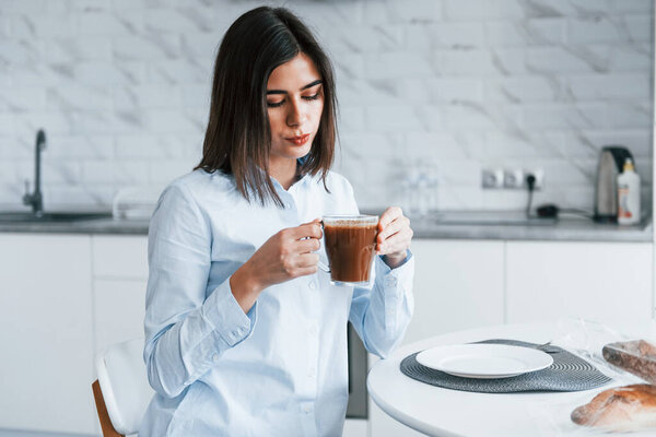 With cup of coffee. Young woman is indoors in room of modern house at daytime.