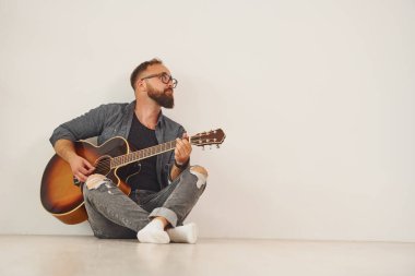 Duvara yaslanarak. Gündelik giysiler içinde ve akustik gitarlı bir adam içeride..