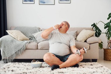 Yemek yiyor ve egzersiz yapıyor. Sıradan giysili komik şişman adam evde..