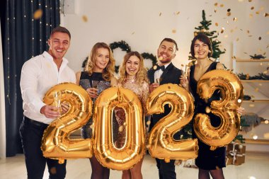 2023 numaralı balonları tutuyorum. Bir grup insan içeride yeni yıl partisi veriyor..