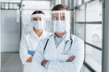 Kollarını kavuşturmuş duruyorsun. Beyaz önlüklü iki doktor klinikte birlikte çalışıyorlar..