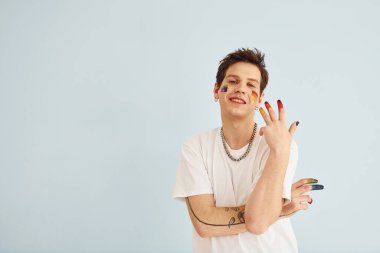 Genç eşcinsel adam stüdyoda duruyor ve kameraya poz veriyor. Çok renkli makyajla boyanmış..