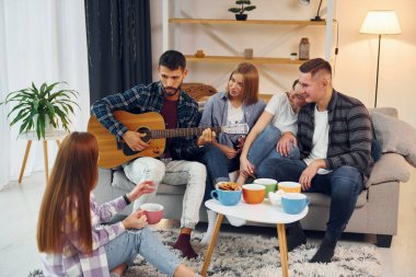 Akustik gitar çalıyor. Bir grup arkadaş içeride parti veriyor..