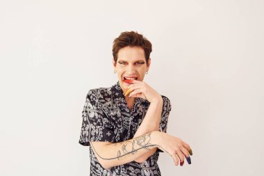 Genç eşcinsel adam stüdyoda duruyor ve kameraya poz veriyor. Yüz parmaklarında çok renkli makyaj malzemeleri var..