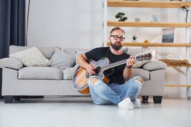 Kanepenin yanında oturuyor. Gündelik giysiler içinde ve akustik gitarlı bir adam içeride..