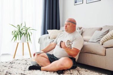 Yemek yiyor ve egzersiz yapıyor. Sıradan giysili komik şişman adam evde..