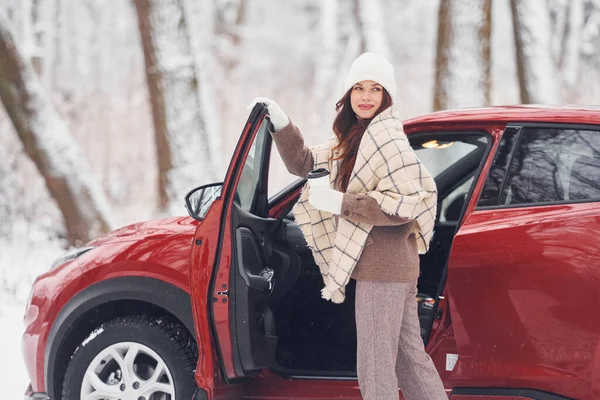 Warm drink in cup. Beautiful young woman is outdoors near her red automobile at winter time.
