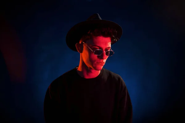In hat and glasses. Neon lighting. Young european man is in the dark studio.