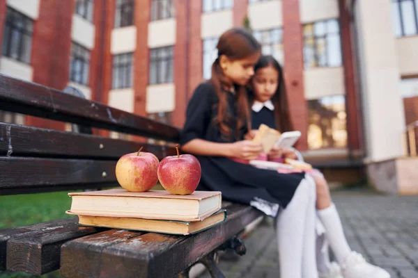 Books and apples. Two schoolgirls is outside together near school building.