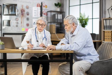 50 yaşındaki kadın doktor, yaşlı hastanın tıbbi muayenesi sırasında panoya yazı yazıyor.