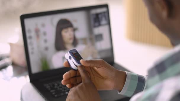 Hombre africano tratando de oxímetro de pulso, escuchando al médico femenino a través de chat de vídeo en línea en la pantalla del ordenador portátil — Vídeos de Stock