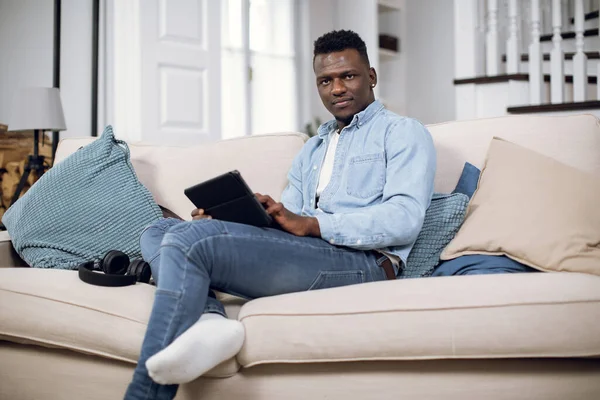 Young african guy sitting on sofa and holding tablet